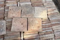 Tomettes carrées beiges et rosées présentées au centre d’une caisse de carreaux rangés sur chant.